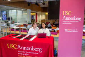 Two USC Annenberg students sitting at a red welcome table with name tags and lanyards, representing the Center for Third Space Thinking in a bright, modern lounge area. A large banner displaying 'USC Annenberg School for Communication and Journalism' is positioned to the right.