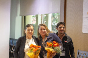 Three individuals smiling while holding vibrant orange floral bouquets, standing indoors near a large window with natural light streaming in. The setting appears warm and celebratory, reflecting an appreciation moment.